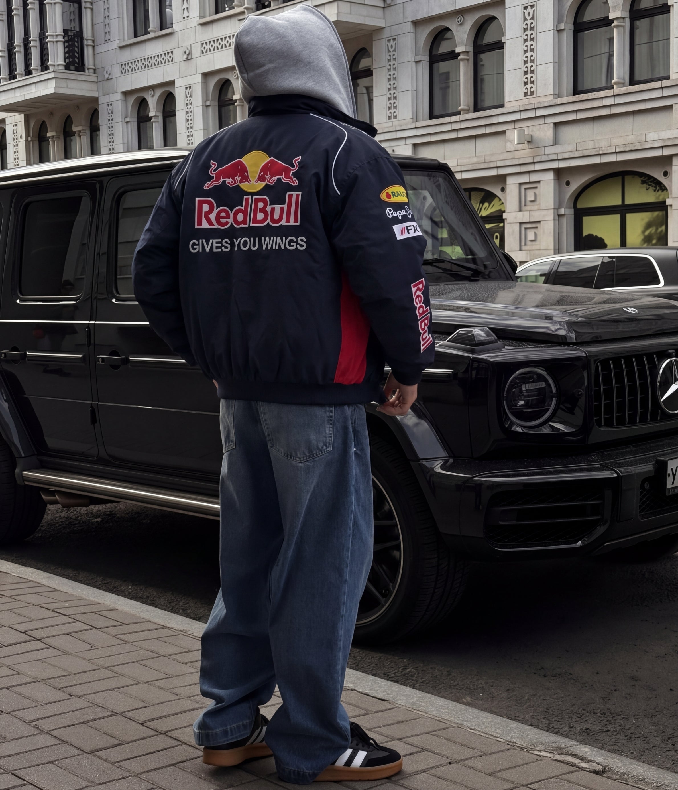Person wearing a Red Bull jacket standing next to a black Mercedes-Benz G-Class in an urban setting.